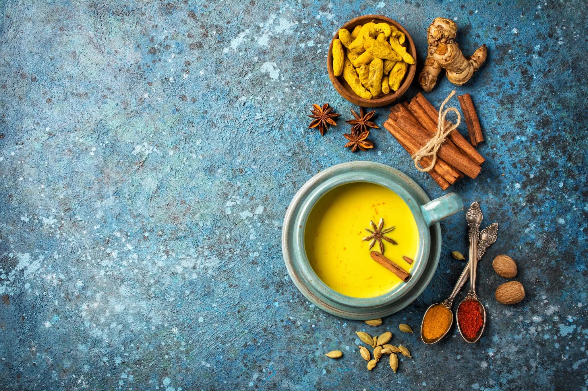 A cup of golden milk with turmeric, cinnamon sticks, star anise, and spices on a textured blue surface