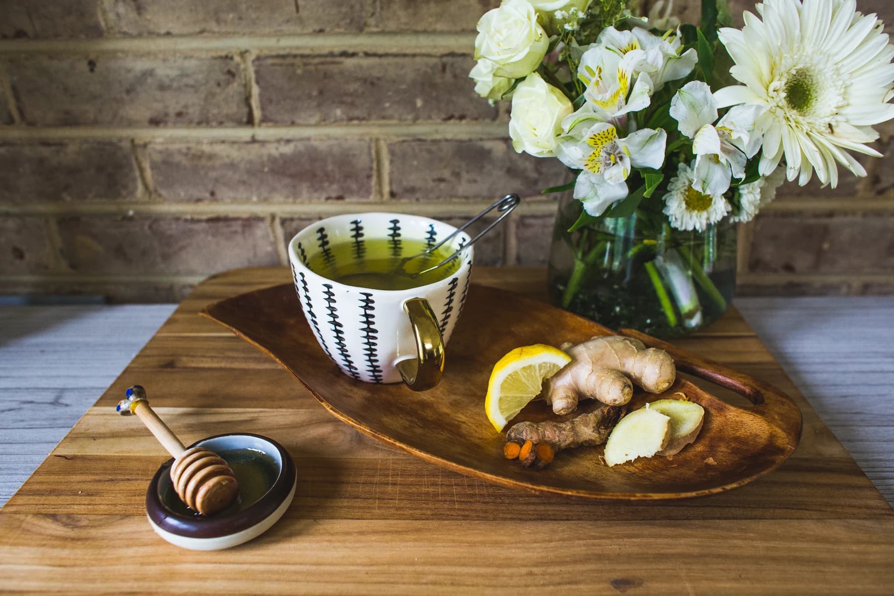 A cup of ginger tea beside fresh ginger, lemon, and white flowers on a wooden tray