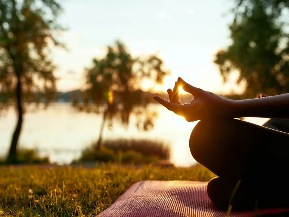 Hands resting in a meditation mudra on a yoga mat beside a lake at sunrise