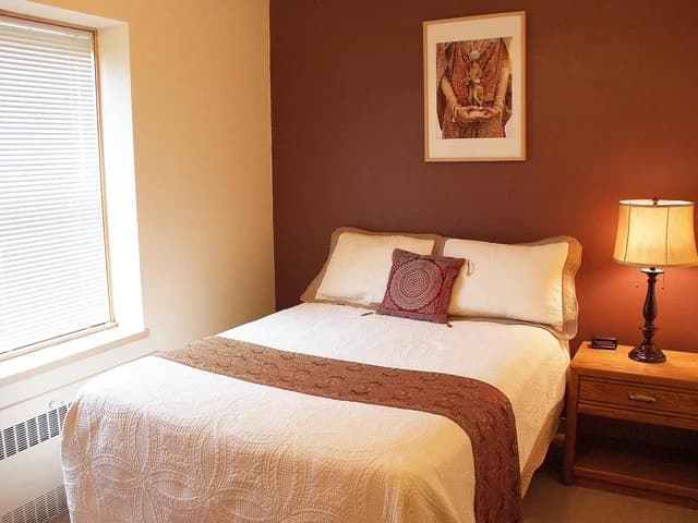 A deluxe suite with a queen bed, a lamp, and a window with blinds