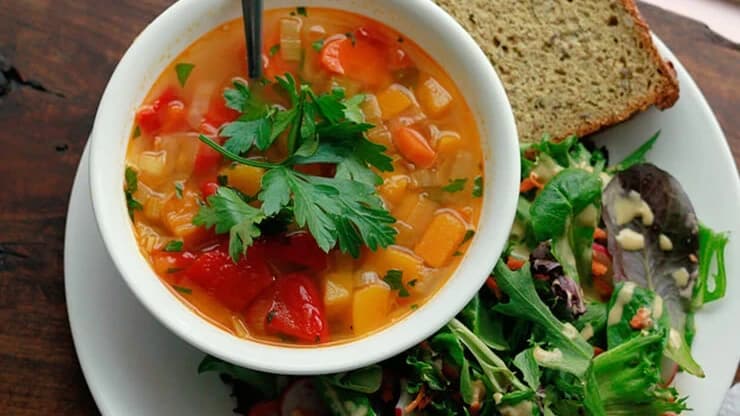 A bowl of vegetable soup with parsley, a slice of bread, and a green salad